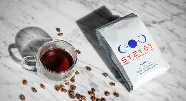 Syzygy coffee bag, a glass of black coffee and roasted coffee beans on a white marble table