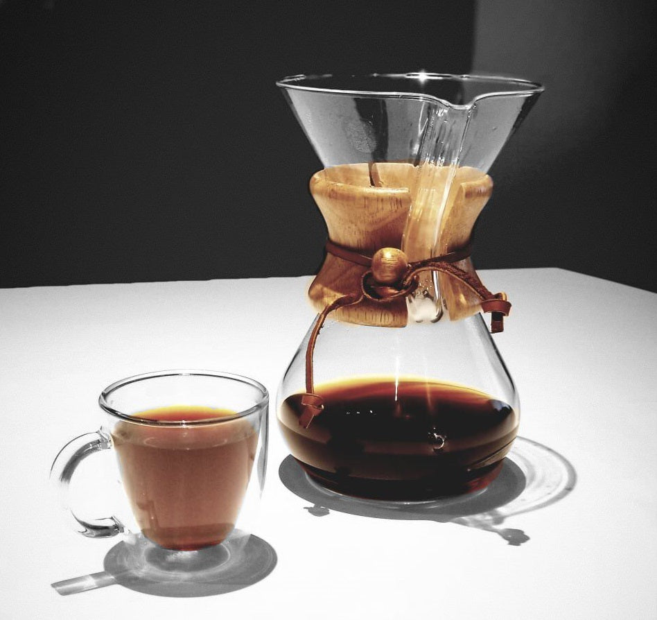 A picture of a chemex coffee maker half filled with coffee alongside a double walled glass coffee mug filled with coffee.