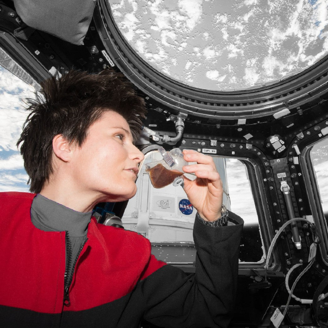 The image shows Italian astronaut Samantha Cristoforetti aboard the International Space Station, looking out the window, while drinking a coffee she brewed using the first espresso maker in space - the ISSPresso. 